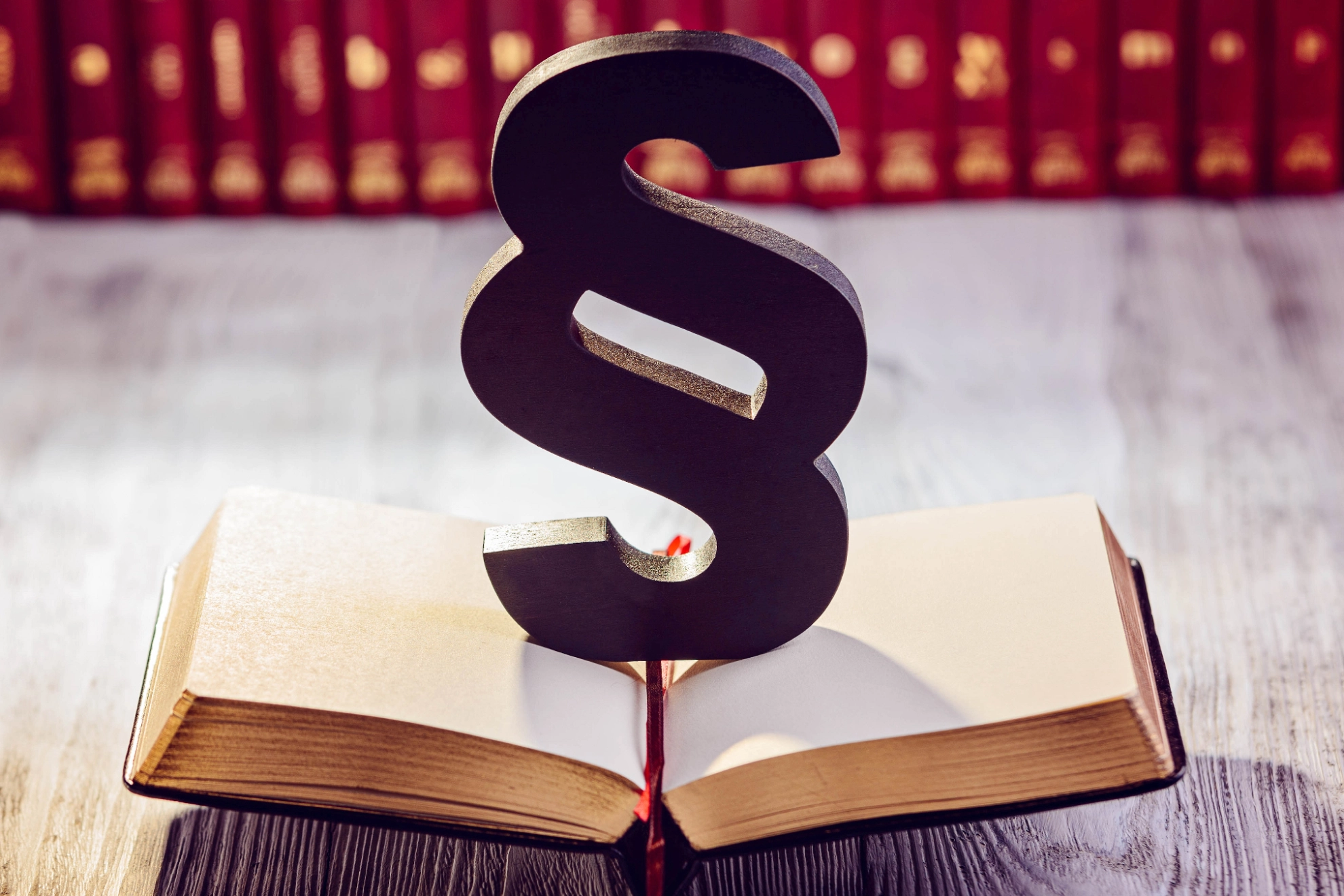 Ein schwarzer Paragraph steht auf einem geöffneten Gesetzbuch, im Hintergrund sind unscharf weitere Bücher zu sehen